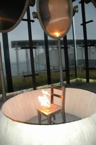 Hanging high above the chair are oval brass mirrors that reflect the light. That's all there is to this memorial, but, in my opinion, no more words are needed. I found it to be a chilling and yet holy place. When I showed this photo to my cousin Kristin Skoglund and her husband, he got it right away. ``The flames are the spirits of those who were killed, rising up away from this place,'' he said. — at Steilneset Memorial in Vardø, Norway.